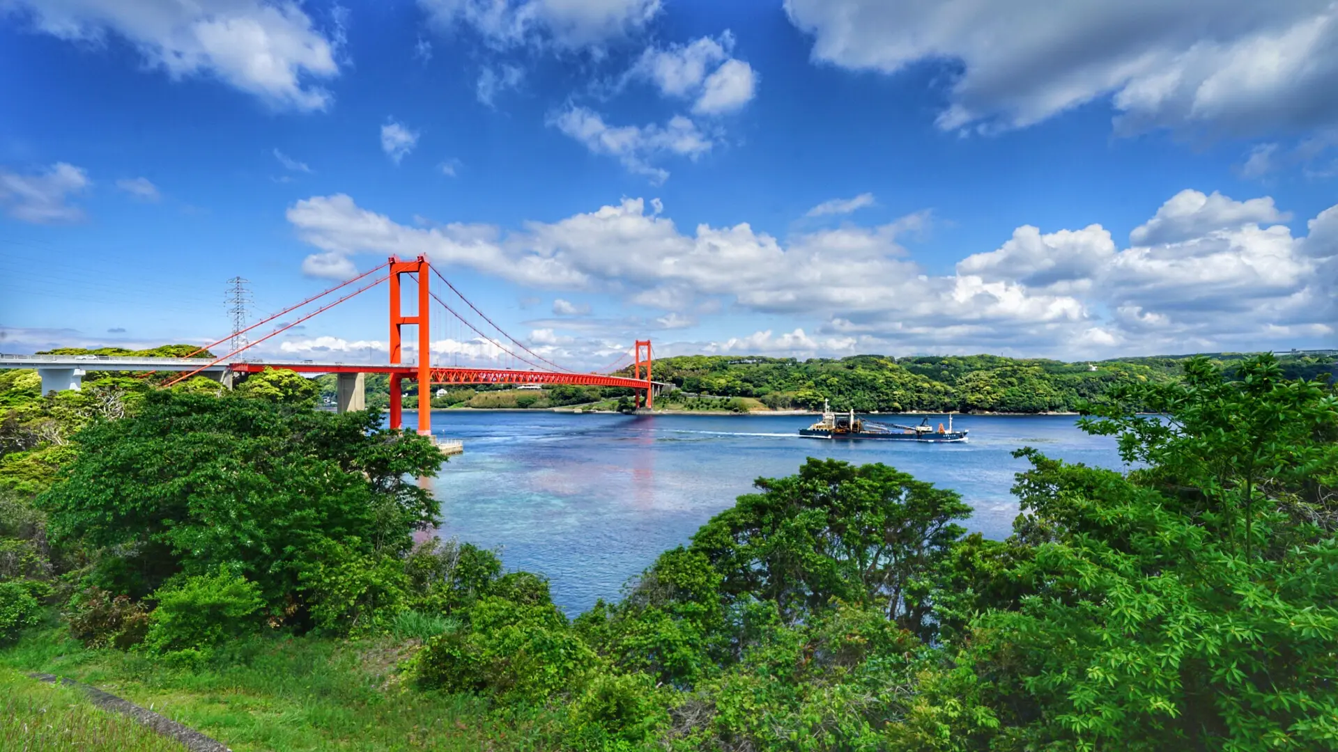 Red suspension bridge spanning a blue river, with green trees on both shores and a boat in the water nearby.