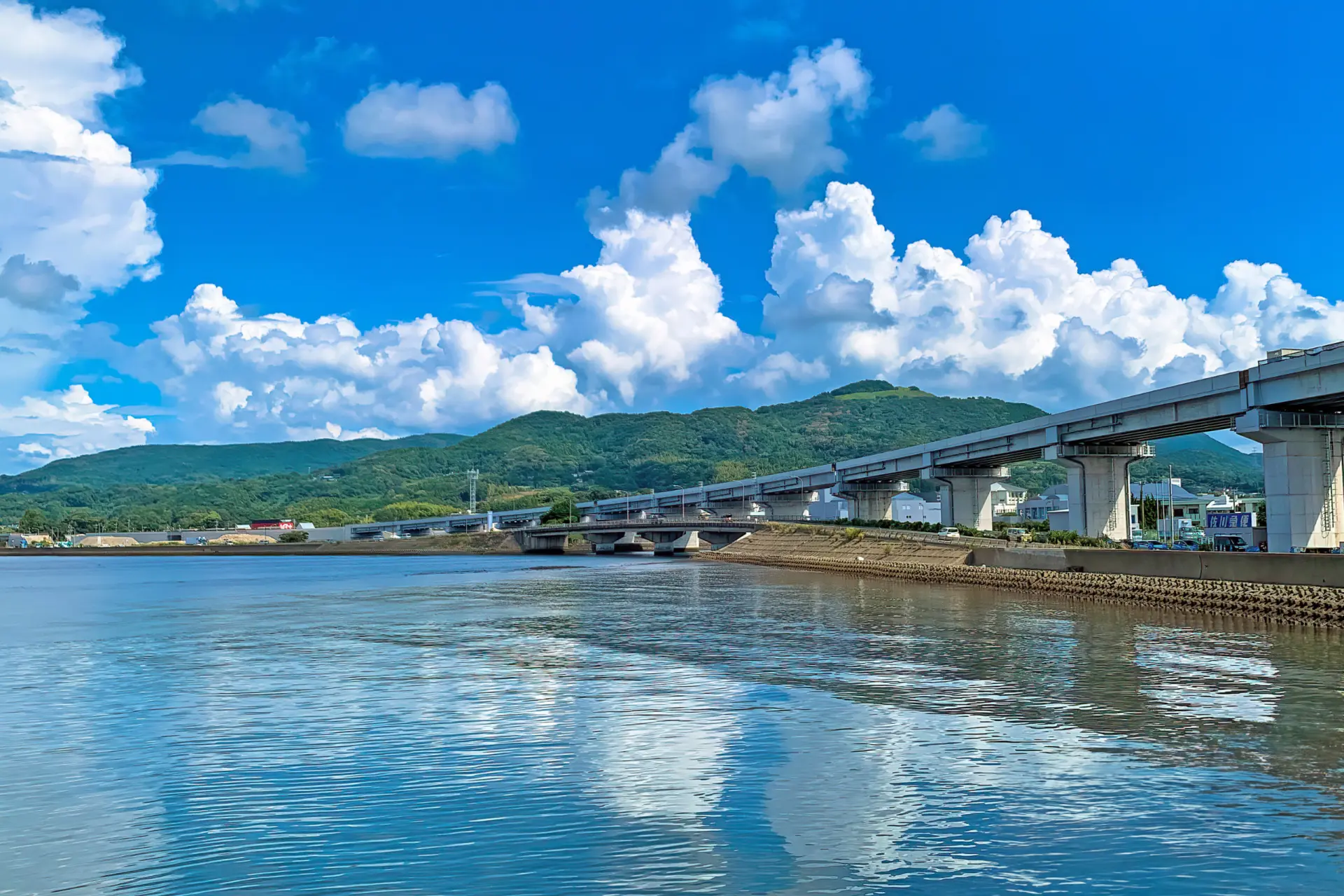 Bridge viaduct stretching over a calm river with green hills in the background under a bright blue sky and fluffy white clouds.